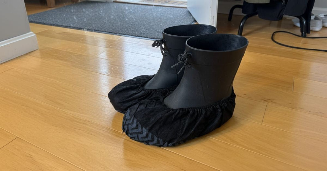 A pair of black rubber boots covered with disposable black shoe covers resting on a light wooden floor in an indoor entryway