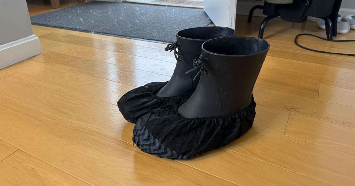 A pair of black rubber boots covered with disposable black shoe covers resting on a light wooden floor in an indoor entryway